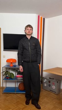 Jauns vīrietis melnā šķūņa jaka un vaļīgās biksēs stāv stūrī istabā ar oranžu lampu, basketbola bumbu, plauktiem un sudraba čemodānu ar uzrakstu Tropicana.