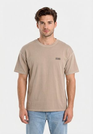 Uomo con capelli castani corti che indossa una T-shirt beige a maniche corte con il logo "American Vintage" e jeans azzurri, in piedi su sfondo bianco.