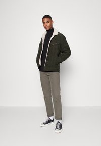 Giacca di velluto a coste verde con colletto in shearling bianco, abbinata a un dolcevita nero, pantaloni grigi chiari e sneakers nere.