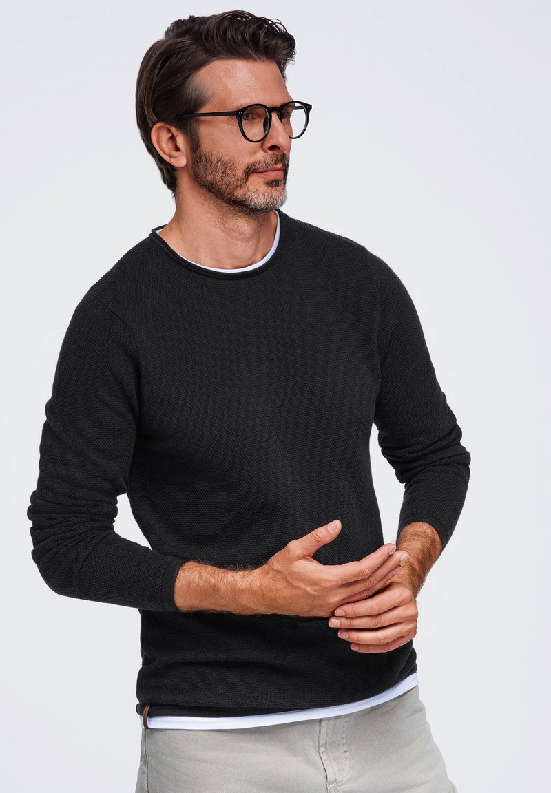 Homme aux cheveux foncés et lunettes, portant un pull noir et un pantalon gris clair sur un fond uni, les mains jointes au niveau de la taille.