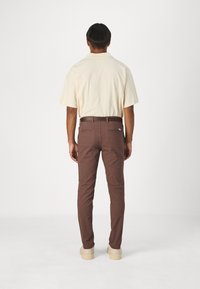 Braune Baumwollhosen im Slim Fit, mit zwei Gesäßtaschen und Gürtelschlaufen, getragen mit einem hellbeigen Kurzarmhemd und beigen Schuhen.