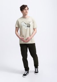 Lichtbeige t-shirt met een bedrukte uil en vleugeldesign; gecombineerd met donkergroene broek. Sneakers met zwart-wit kleurenpalet.