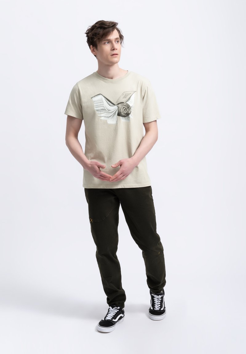 Lichtbeige t-shirt met een bedrukte uil en vleugeldesign; gecombineerd met donkergroene broek. Sneakers met zwart-wit kleurenpalet.