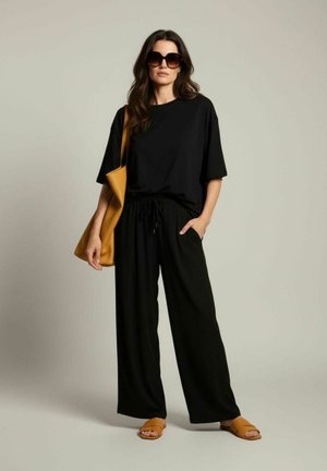 Femme portant une tenue noire ample, des lunettes de soleil oversize, des sandales marron à enfiler et portant un grand sac à épaule marron sur fond uni.