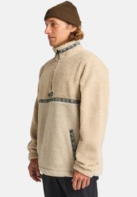 Beige fleece pullover med halv-lynlås krave, der har dekorative blå og sorte kanter langs brystet og lommen for ekstra detalje.