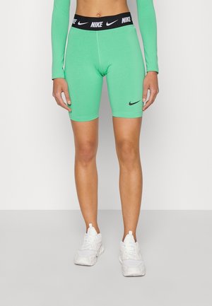 Person iført grønne Nike cykelshorts med sort taljebånd og hvide sneakers, stående mod en ensfarvet lys baggrund.