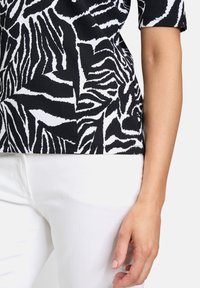 Top a righe zebra bianco e nero con texture a costine, dotato di maniche corte; abbinato a pantaloni bianchi. Mano visibile a destra.