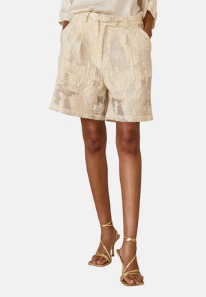 Donna che indossa pantaloncini beige testurizzati al ginocchio con cintura e sandali dorati con tacco alto e cinturini, mani in tasca.