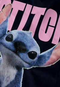 Niebieska pluszowa postać z dużymi czarnymi oczami i różowymi uszami na ciemnogranatowym tle, obok dużego, jasnoróżowego napisu "STITCH".