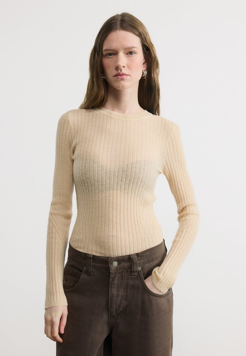 Jeune femme aux longs cheveux bruns portant un pull en maille côtelée beige et un pantalon marron foncé, une main dans la poche, sur un fond blanc uni.