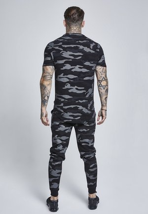 Mand med tatoveringer står med fronten væk iført sort og gråt camouflage kortærmet skjorte og matchende bukser med sorte sneakers på en ensfarvet baggrund.