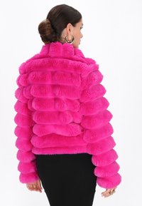 Vrouw met een knot, gekleed in grote gouden huggies en een felroze, textuurachtige bontjas, met de rug naar ons toe gekeerd en een zwarte rok.