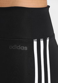 Czarne legginsy z wysokim stanem, z trzema białymi pionowymi paskami i szarym logo Adidasa po lewej stronie. Gładka tekstura.