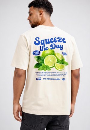 Man wearing beige t-shirt with back graphic of limes, mint leaves, and text "Squeeze the Day" promoting juicy mojitos.