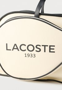 Kremowa teksturowana torba na ramię z czarnymi skórzanymi akcentami i uchwytami. Wyraźne oznaczenie "LACOSTE 1933" w pogrubionych, ciemnych literach.