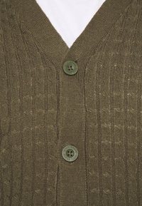 Kardigan oliwkowozielony z fakturowanym wzorem dzianiny, z głębokim dekoltem w kształcie litery V, dwoma zielonymi guzikami i warstwowym wyglądem na białej koszuli.