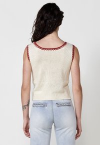 Top de punto sin mangas en color crema con un acabado texturizado, que presenta un escote y sisas en forma de ondulación color burdeos oscuro, combinado con unos jeans de color azul claro.