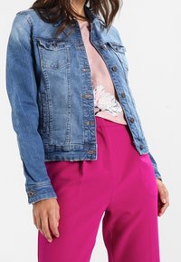 Chaqueta vaquera en azul claro, con dos bolsillos en el pecho, botones plateados y una textura ligeramente desgastada, combinada con una camiseta y pantalones rosas.