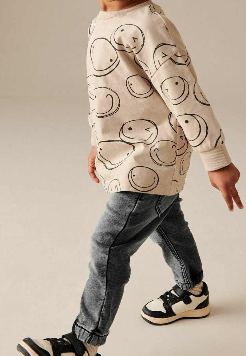 Enfant portant un sweat-shirt beige avec des visages souriants en contour noir, un pantalon de jogging gris et des baskets noir et blanc, marchant devant un fond uni.