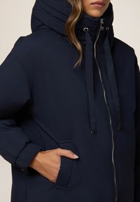 Mørkeblå jakke med høj krave og justerbare snore, med frontlynlås, sidelommer og et glat, let stof.