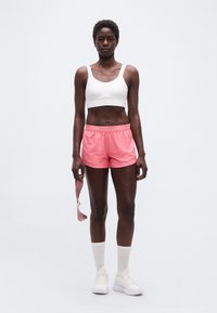 Γυναίκα που στέκεται φορώντας λευκό αθλητικό σουτιέν, ροζ αθλητικά σορτς, λευκές κάλτσες και αθλητικά παπούτσια, κρατώντας μια ροζ λάστιχο αντίστασης σε απλό φόντο.