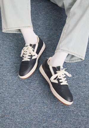 Mornarski usnjeni športniki z belimi detajli in vezalkami; gumijast podplat z rjavimi poudarki; postavljeno proti modri preprogi.