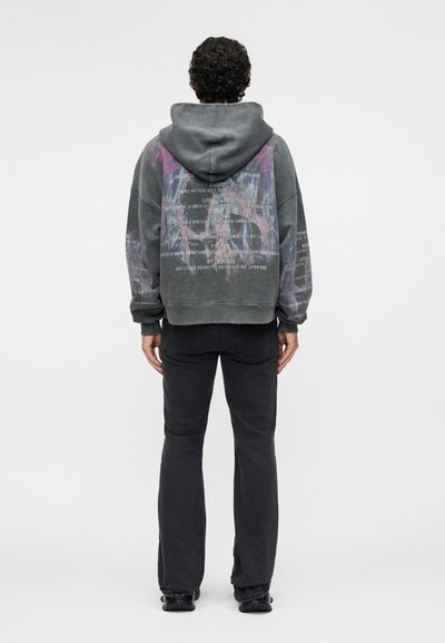 Osoba s kučeravými vlasmi stojí chrbtom, má na sebe vybledlú sivú mikinu s kapucňou s textom a farebnými grafickými detailmi na chrbte a rukávoch, čierne rifle.