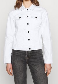 Mujer vestida con una chaqueta de mezclilla blanca ajustada con botones negros y jeans gris oscuro, de pie contra un fondo claro y liso.