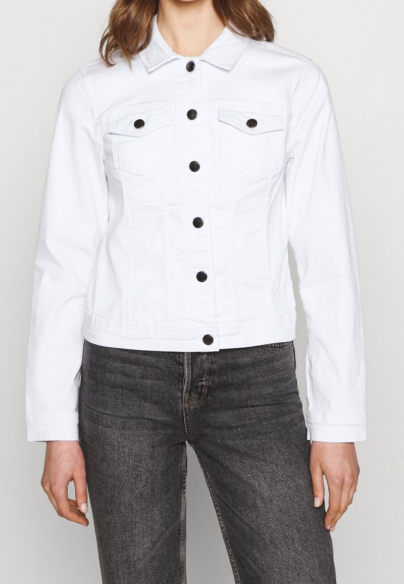 Mujer vestida con una chaqueta de mezclilla blanca ajustada con botones negros y jeans gris oscuro, de pie contra un fondo claro y liso.