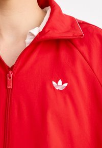 Czerwona kurtka zapinana na zamek z usztywnianym kołnierzem i białym logo. Wykonana z gładkiej tkaniny o lekkiej teksturze, z centralnym zamkiem błyskawicznym.