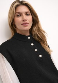 Black sleeveless sweater with a high collar and five large, round buttons at the front. The fabric appears soft and textured.