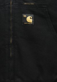 Črna jakna z zlatim zadrgo, oznako "Anniversary Edition" in kvadratno nalepko z "50" ter logotipom Carhartt v zlati barvi.