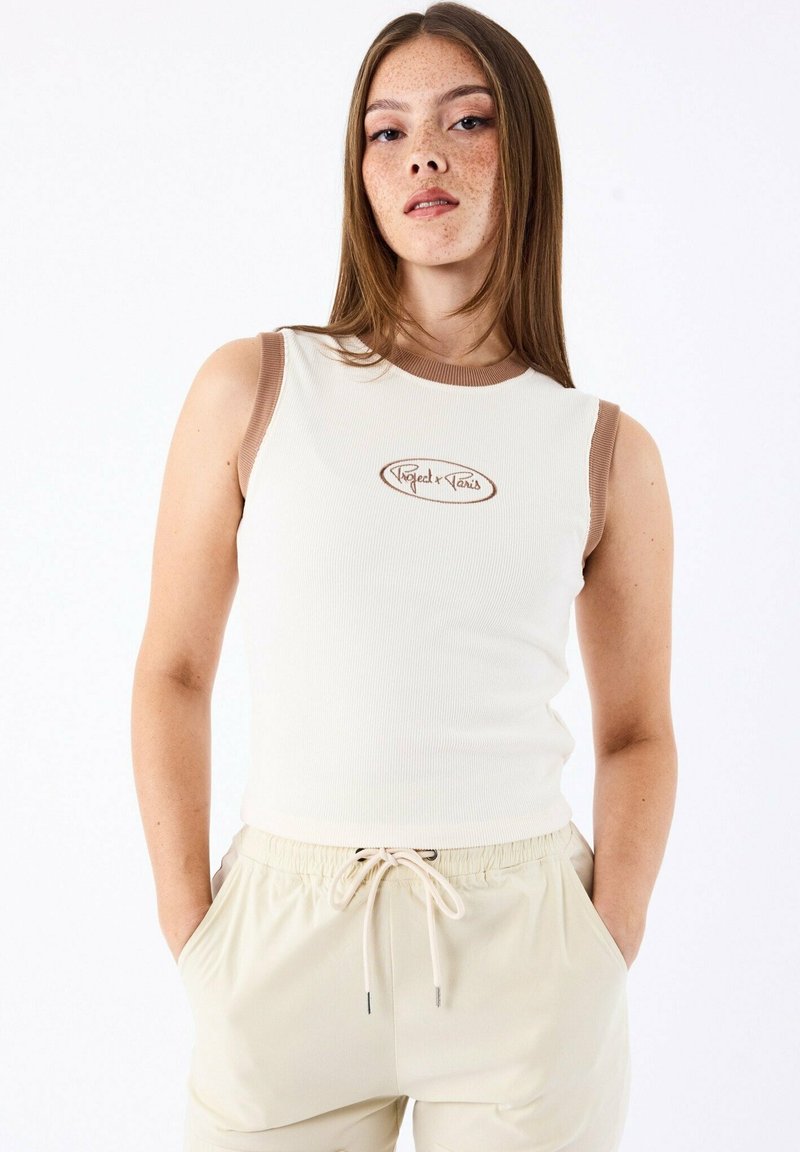 Camiseta de tirantes acanalada en color blanco roto con detalles en marrón y logotipo bordado. Presenta tirantes anchos y una silueta ajustada. Combinada con pantalones beige.