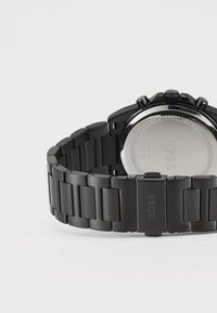Montre en métal noir avec un bracelet à maillons, un boîtier rond et un dos en acier inoxydable. Présente un texte gravé pour la résistance à l'eau et le logo de la marque.