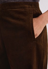 Bruine corduroy broek met verticale ribbels, voorzien van zijzakken en een ontspannen pasvorm. Close-up van de stof en het zakgebied.
