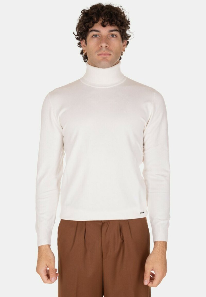 Maglione bianco a collo alto con texture a costine, maniche lunghe e design aderente. Abbinato a pantaloni marroni, con un minimo dettaglio di branding visibile.