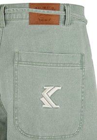 Un pantalon en denim vert clair présente une poche arrière avec un logo brodé blanc et une étiquette en cuir brun avec des détails de la marque.