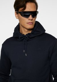 Marineblå hættebluse med snoretræk i kraven, prydet med et struktureret logo på brystet. Sorte sporty solbriller fuldender looket.