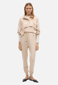 Sudadera con capucha beige con un bolsillo frontal, combinada con joggers ajustados a juego. Tejido suave, diseño minimalista y cordón ajustable.