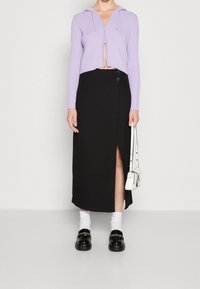 Avkortad lila hoodie med dragkedja och en svart omlottkjol med sidofodral och knappdetalj. Buren med vita strumpor och svarta skor.