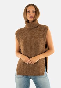 Femme portant un pull en maille sans manches de couleur marron sur un jean bleu clair, debout avec les mains jointes devant elle.