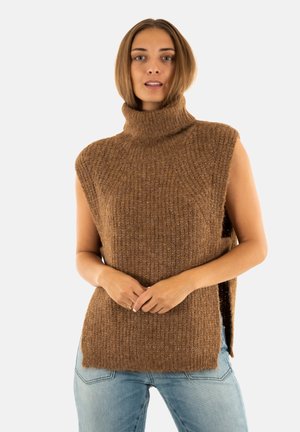 Donna che indossa un maglione senza maniche a collo alto color marrone sopra dei jeans azzurri chiari, in piedi con le mani unite davanti.