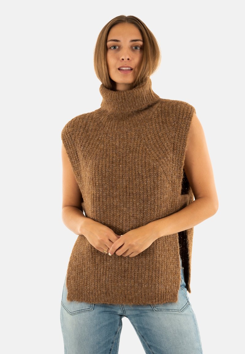 Femme portant un pull en maille sans manches de couleur marron sur un jean bleu clair, debout avec les mains jointes devant elle.