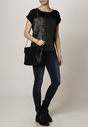 Kobieta ubrana w czarną i srebrną bluzkę z krótkim rękawem, ciemne obcisłe dżinsy, czarne botki za kostkę, trzymająca czarną skórzaną torebkę na pasku przewieszoną przez ramię.