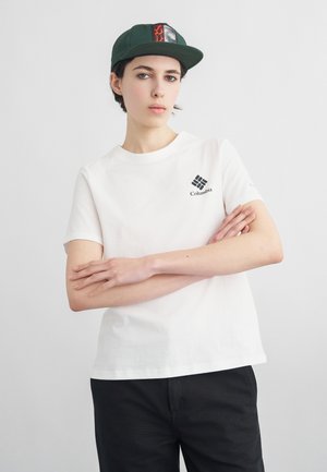 Mladá osoba v bielej tričku Columbia a zelenej šiltovke so založenými rukami na jednoduchom svetlom pozadí.