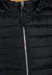 Veste matelassée noire avec une fermeture éclair argentée brillante, texture matelassée, col haut et une boucle en tissu au cou pour un rangement facile.