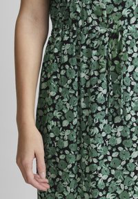 Tumma vihreä kukkamekko, jossa valkoisia kukkia. Ominaisuuksiin kuuluu laskostettu vyötärö ja solmittava yksityiskohta. Materiaali vaikuttaa pehmeältä ja siinä on teksturoidun näköinen pinta.