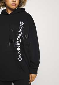 Felpa nera con cappuccio caratterizzata da un logo Calvin Klein Jeans bianco, tessuto liscio, vestibilità oversize e dettagli a contrasto con un nastro nero sul davanti.