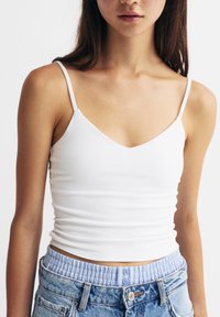 White fitted tank top with thin straps, featuring a deep V-neckline. Worn with high-waisted blue denim shorts with a gathered waistband.