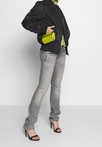 Schwarze Bomberjacke, grünes gemustertes Oberteil, graue Jeans, schwarze Absatzsandalen und eine kleine gr üne Handtasche in der Hand.